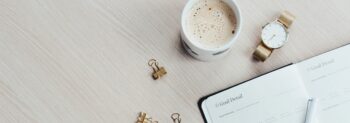 Looking down on a cup of coffee, a watch, and a diary, which are sitting on a desk.